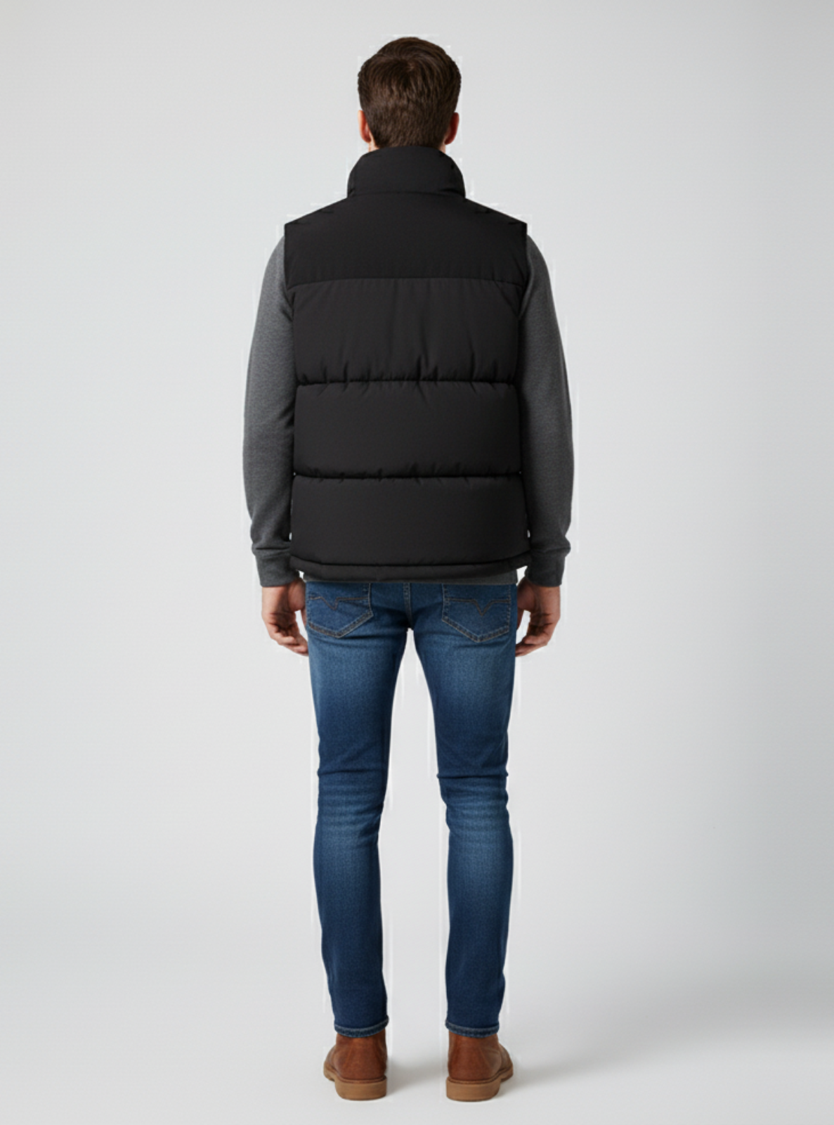 Man wearing a black puffer vest over a gray shirt with blue jeans on a white background