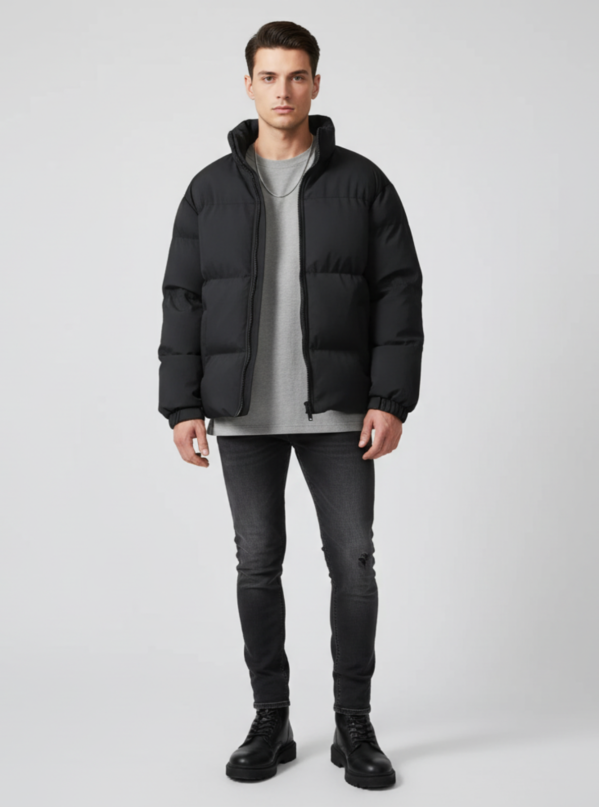 Man wearing a black puffer jacket, gray sweater, black jeans, and black boots on a light gray background