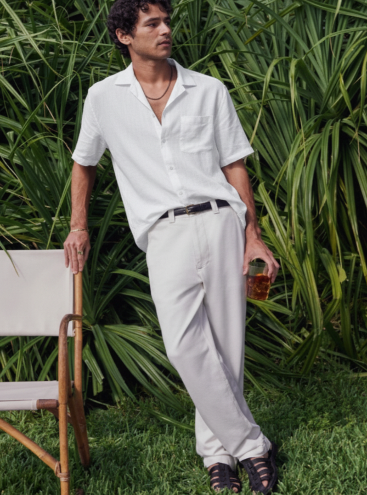 Man in white shirt and light pants standing outdoors with greenery in the background