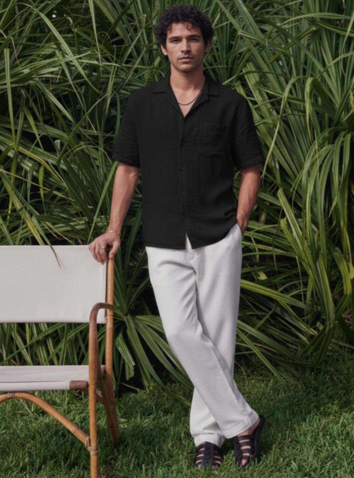Man in black shirt and white pants standing in a grassy area with greenery.