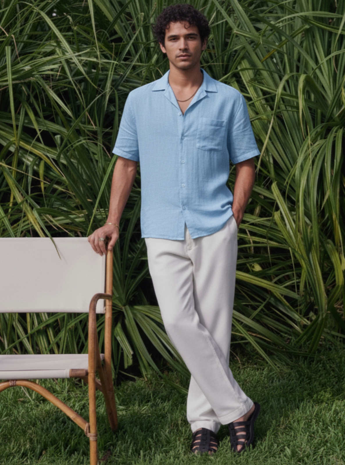 Man in a light blue shirt and white pants standing in a grassy area with greenery.