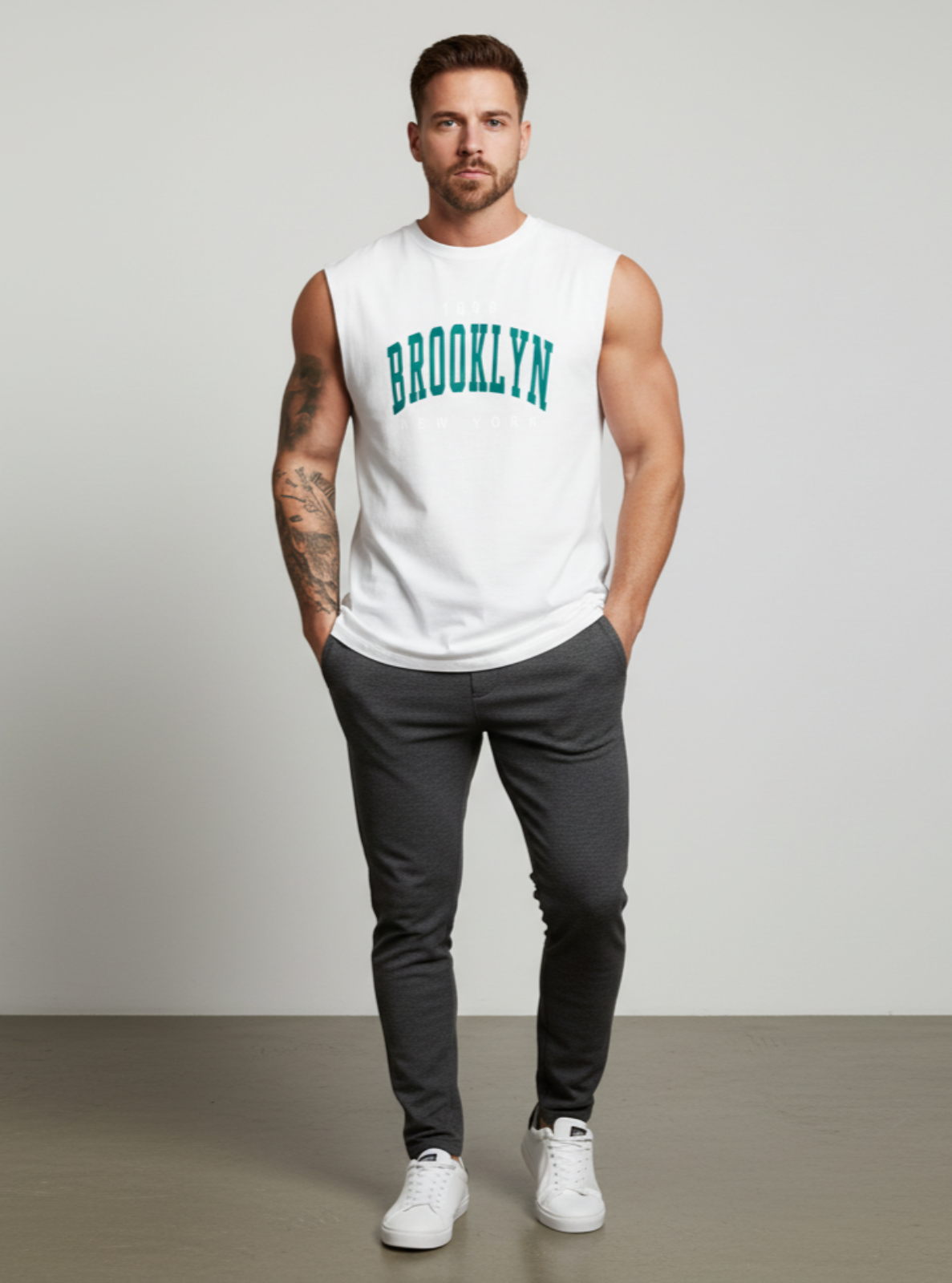 Man wearing a white sleeveless shirt with 'Brooklyn' text, dark pants, and white sneakers on a plain background