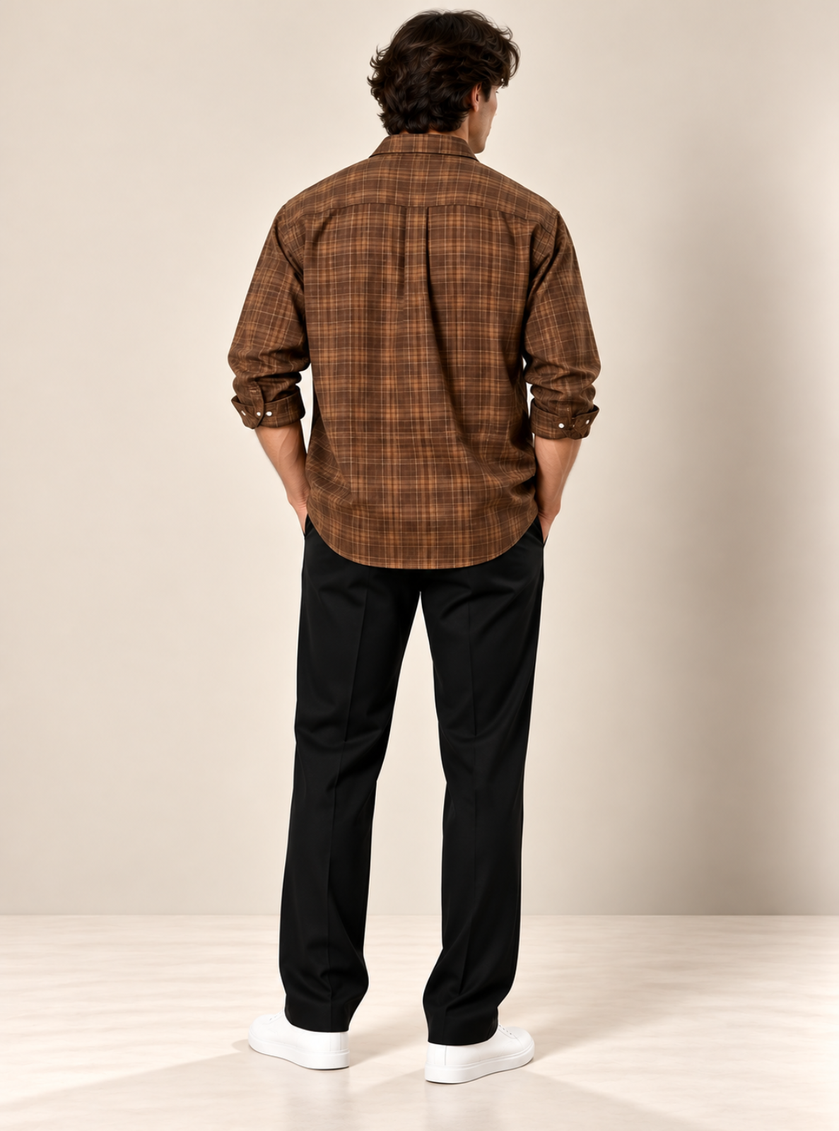 A man viewed from behind wearing a brown and black plaid button-down shirt, black trousers, and white sneakers, standing against a neutral beige studio background.