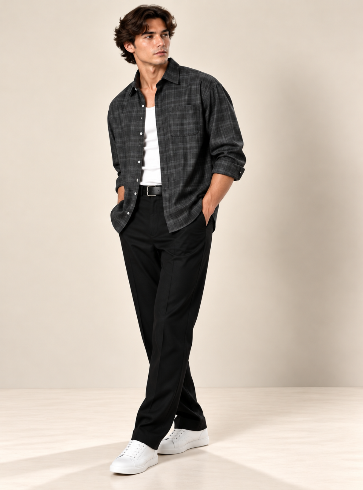 A man modeling a dark gray and black plaid button-down shirt over a white t-shirt, paired with black trousers and white sneakers, standing against a neutral beige studio background.
