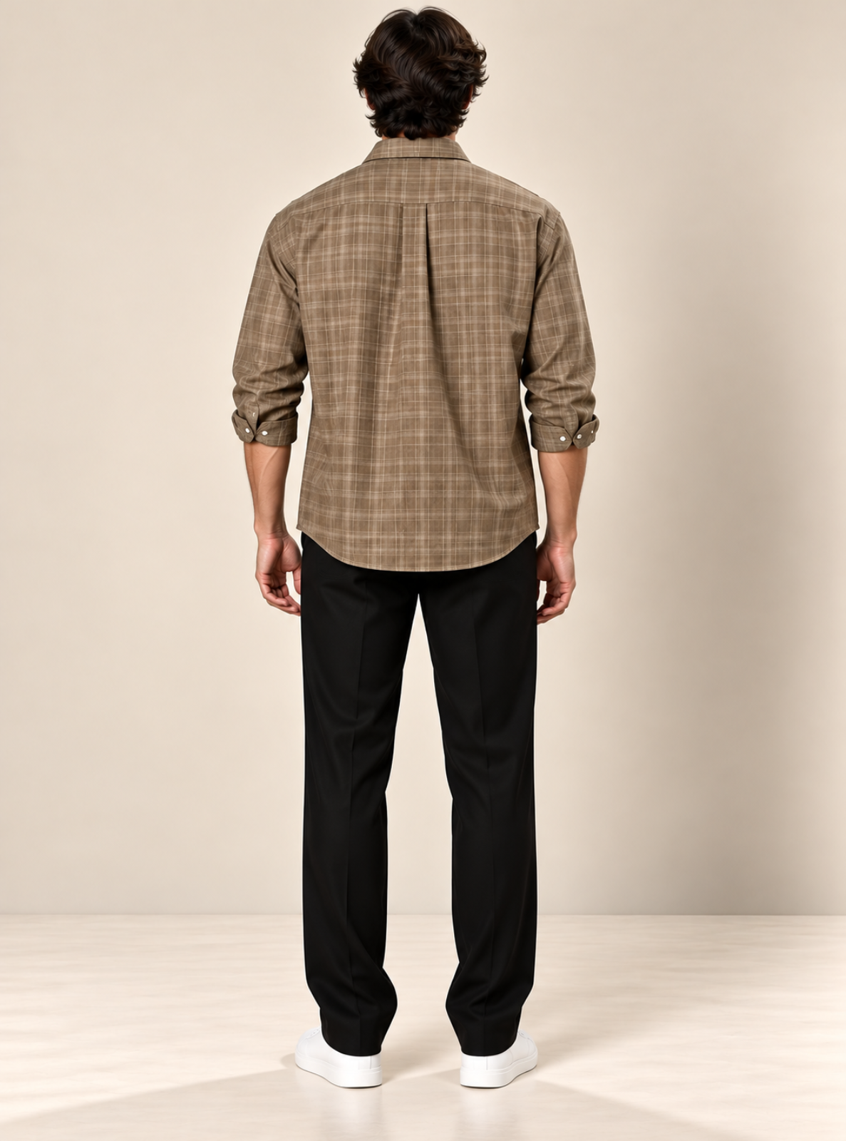 A man viewed from behind wearing a brown and beige plaid button-down shirt, black trousers, and white sneakers, standing against a neutral beige studio background.