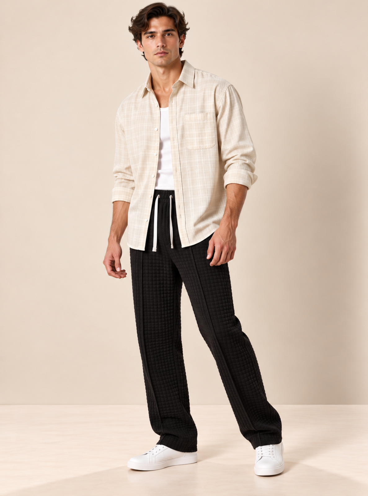 A male model stands in a studio wearing a light beige plaid overshirt over a white shirt, black textured trousers, and white sneakers against a warm beige backdrop.