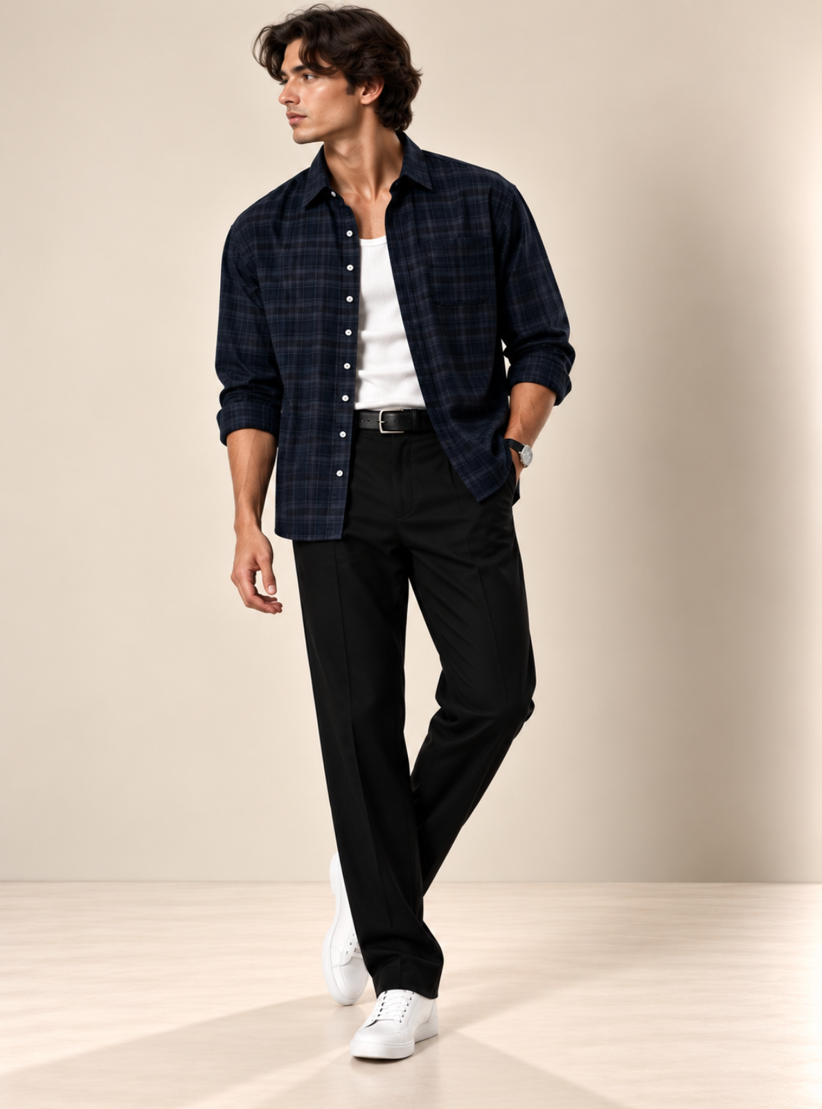 A man modeling a dark navy plaid button-down shirt over a white t-shirt, paired with black trousers and white sneakers, in a studio setting with a beige background.