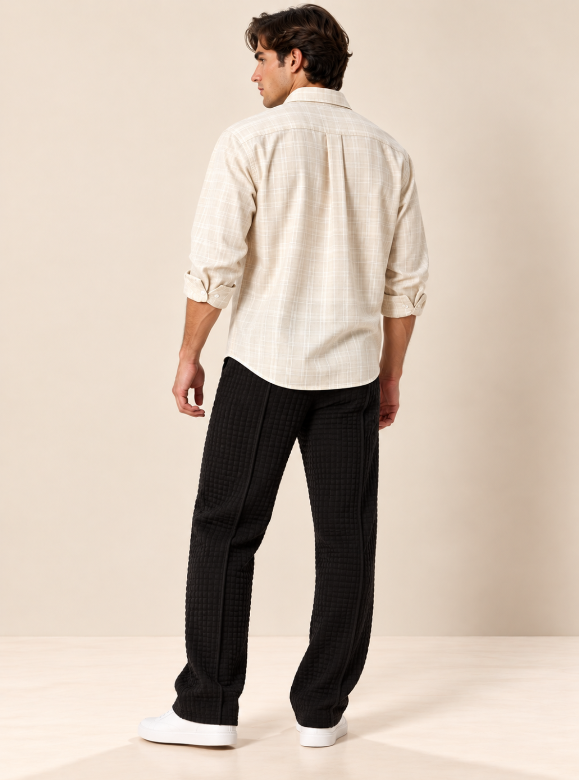 A man shown from the back wearing a cream-colored textured button-down shirt and black textured trousers, standing against a warm beige studio background.