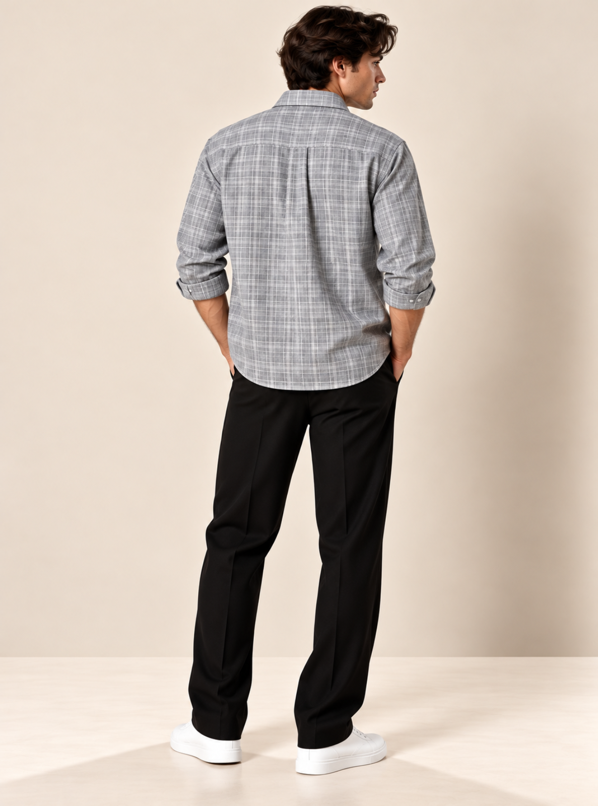 A man is shown from the back wearing a gray and white plaid button-down shirt, black trousers, and white sneakers, standing against a solid beige studio background.
