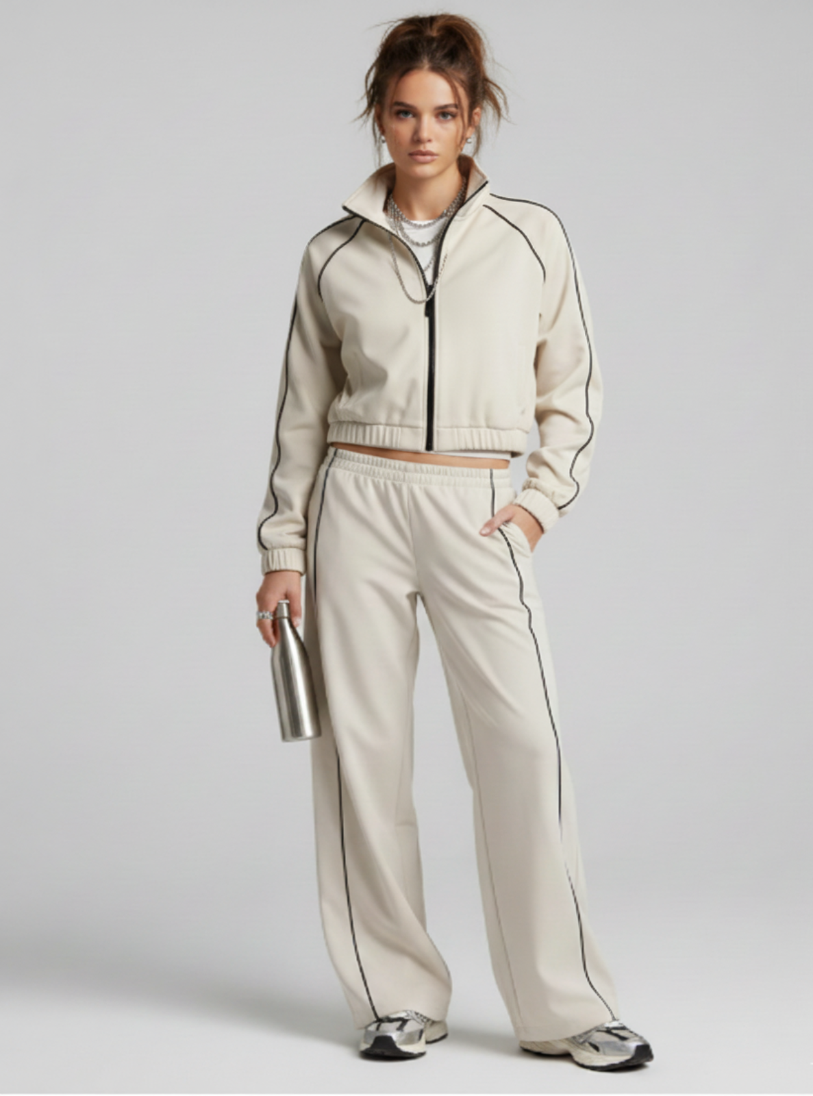 Woman wearing a beige tracksuit holding a silver clutch against a light gray background