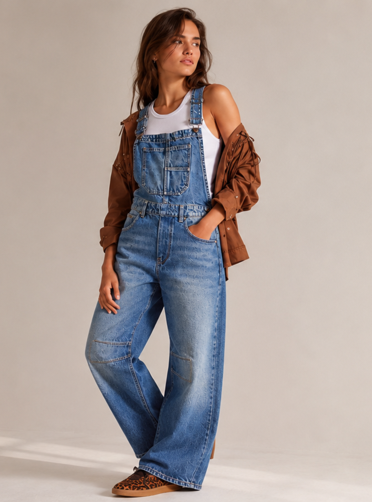 A woman modeling blue denim overalls with a white tank top, brown jacket, and brown shoes, standing in a minimalist studio with warm sunlight and shadows.