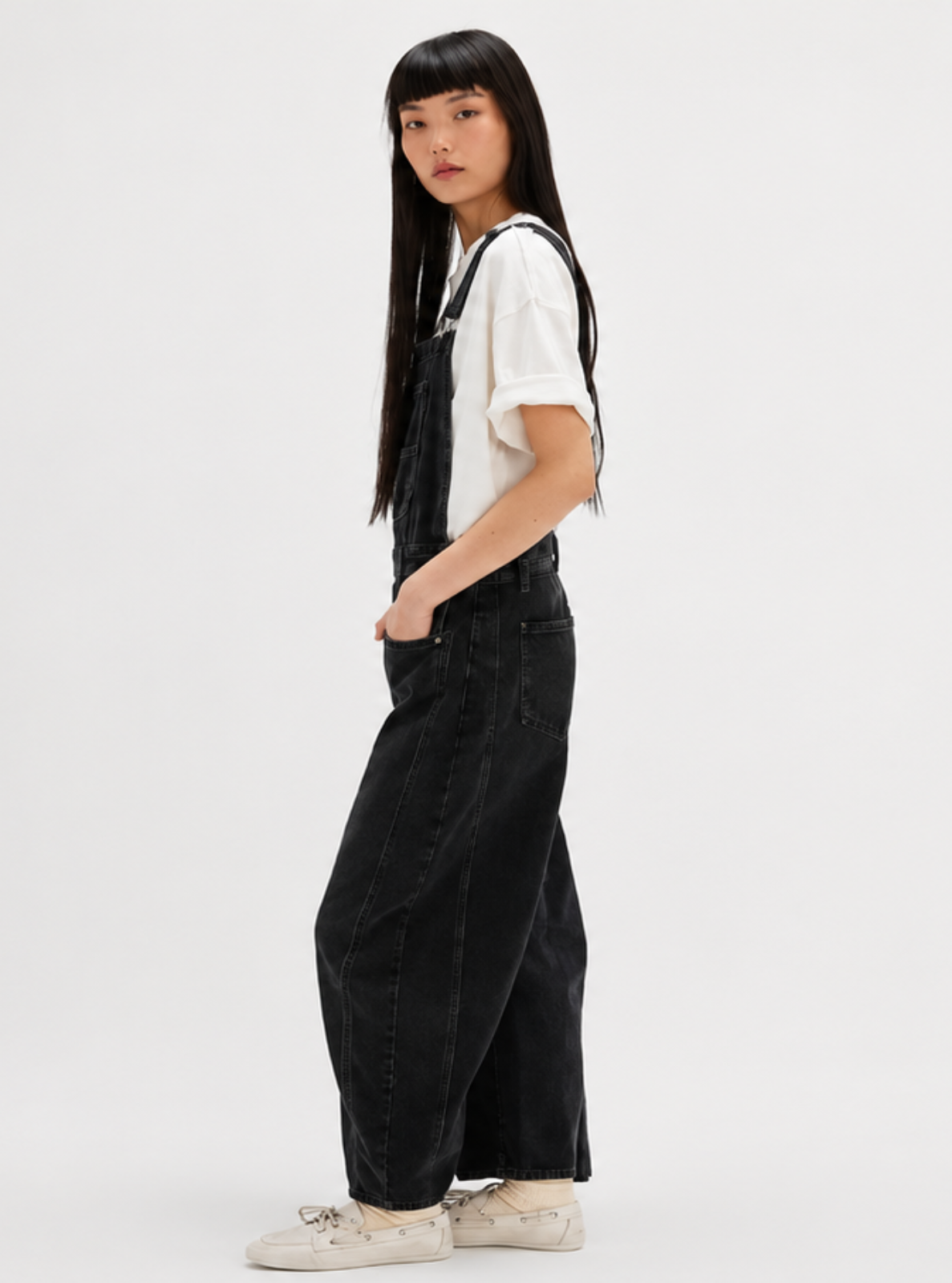 A model stands in profile wearing a white short-sleeve t-shirt and black wide-leg corduroy overalls, paired with white sneakers, against a plain white studio background.