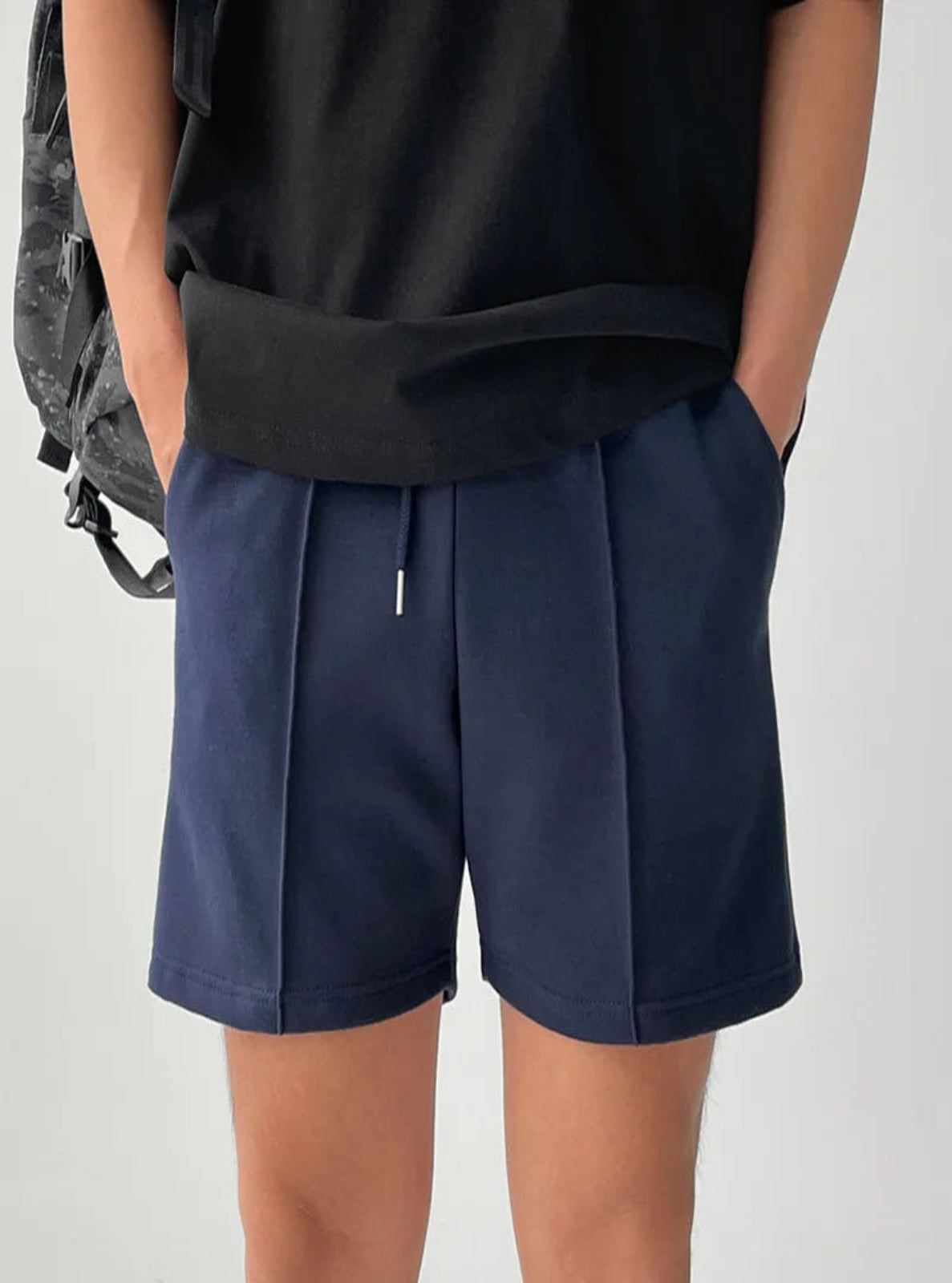 Person wearing navy shorts and a black top with a gray backpack on a light gray background