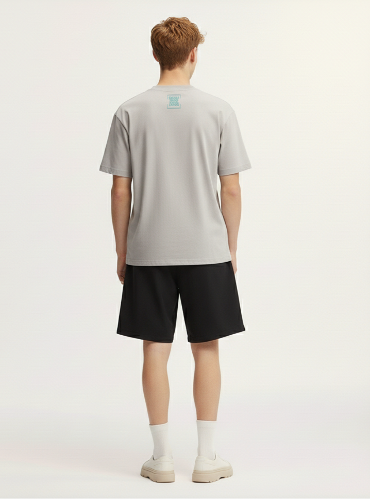 Person wearing a light gray t-shirt with a logo on the back and black shorts against a white background