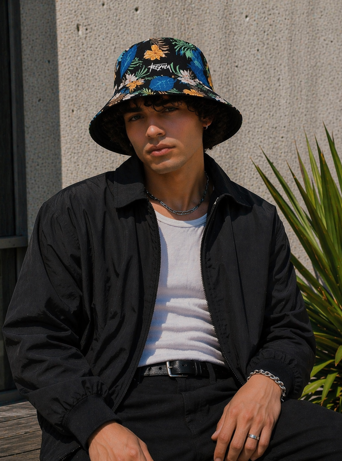 Person wearing a floral bucket hat and black jacket sitting against a concrete wall with a plant.