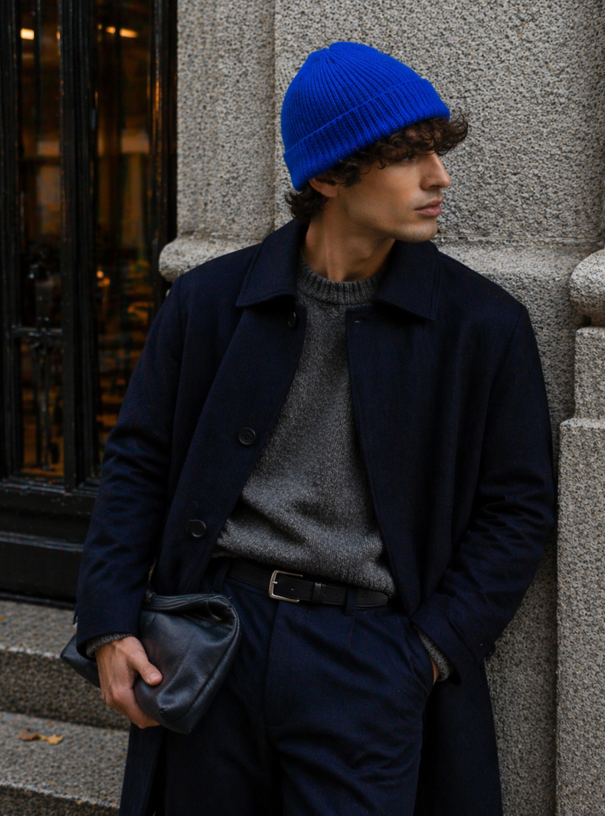 Person wearing a blue beanie and dark coat leaning against a building.