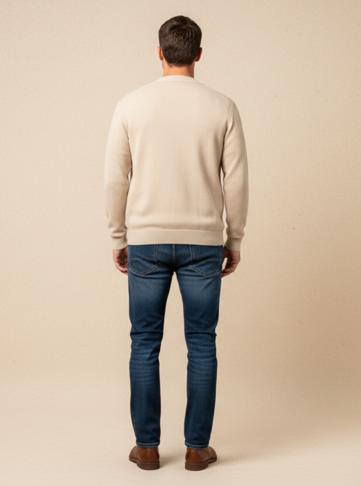 Man wearing a beige sweater and blue jeans standing against a beige background