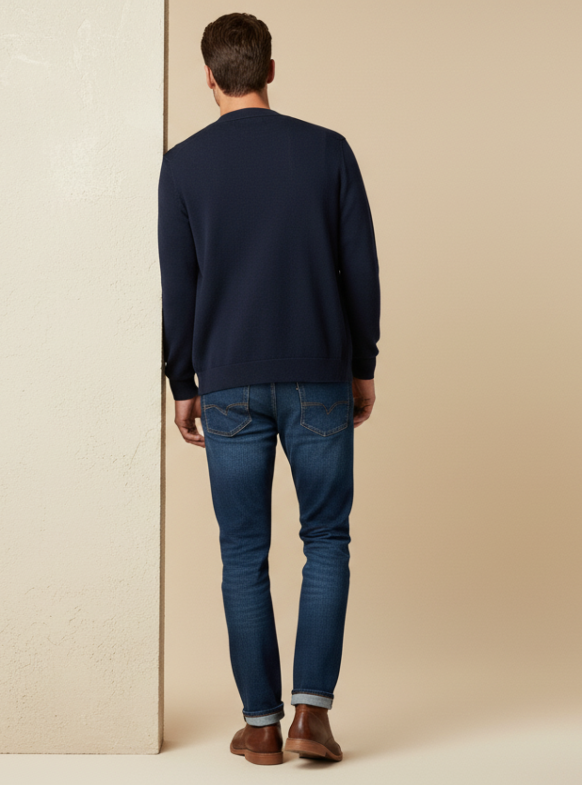 Man wearing a navy sweater and blue jeans standing against a beige wall.
