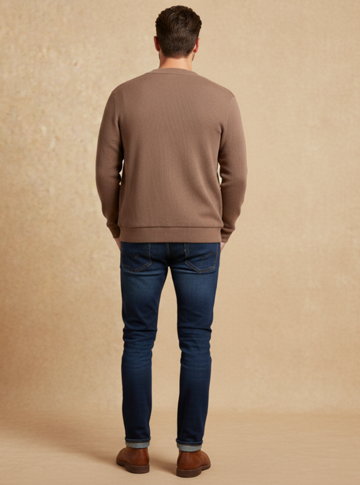 Man wearing a brown sweater and blue jeans standing against a beige background