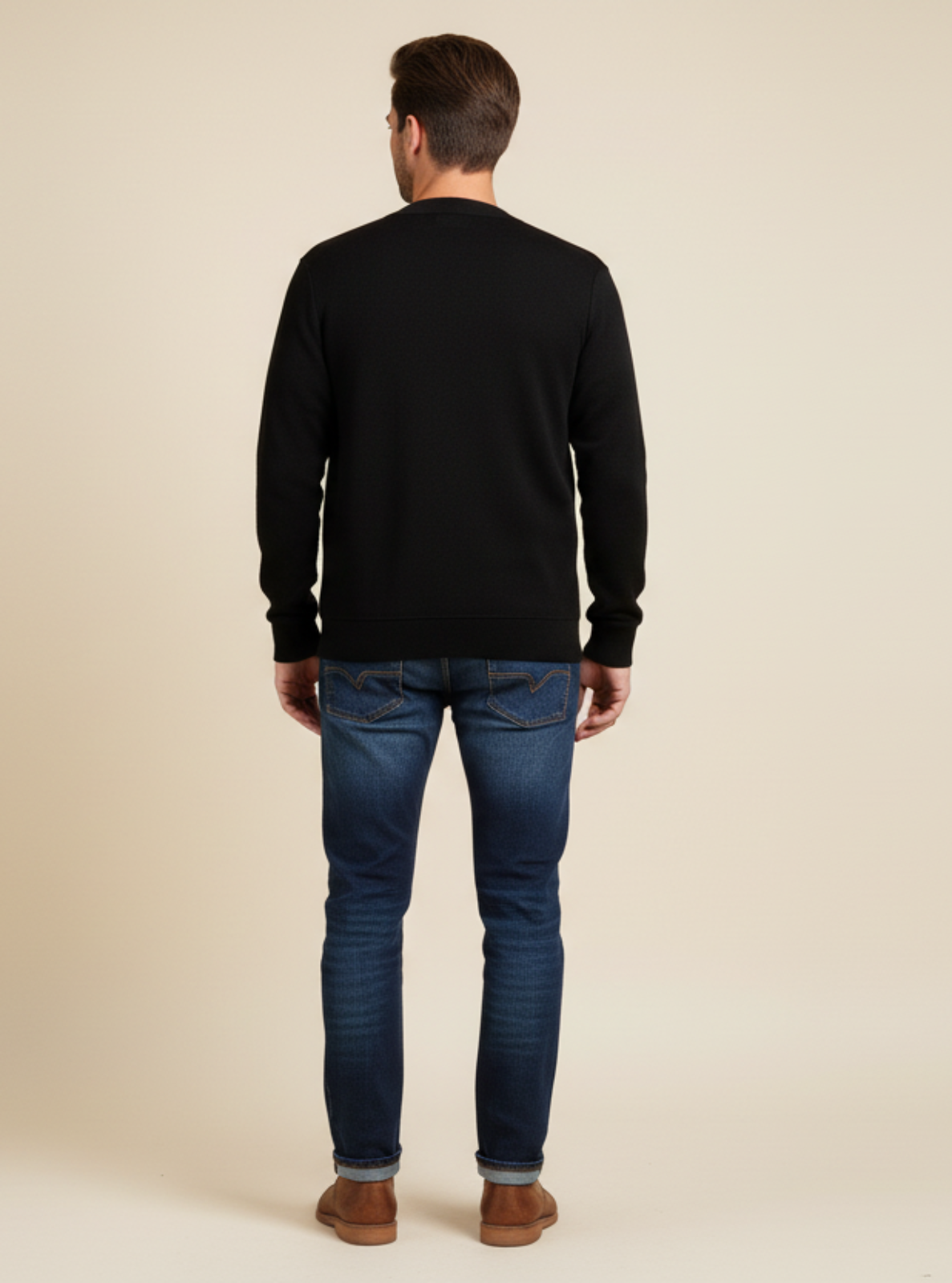 Man wearing a black sweater and blue jeans standing against a beige background