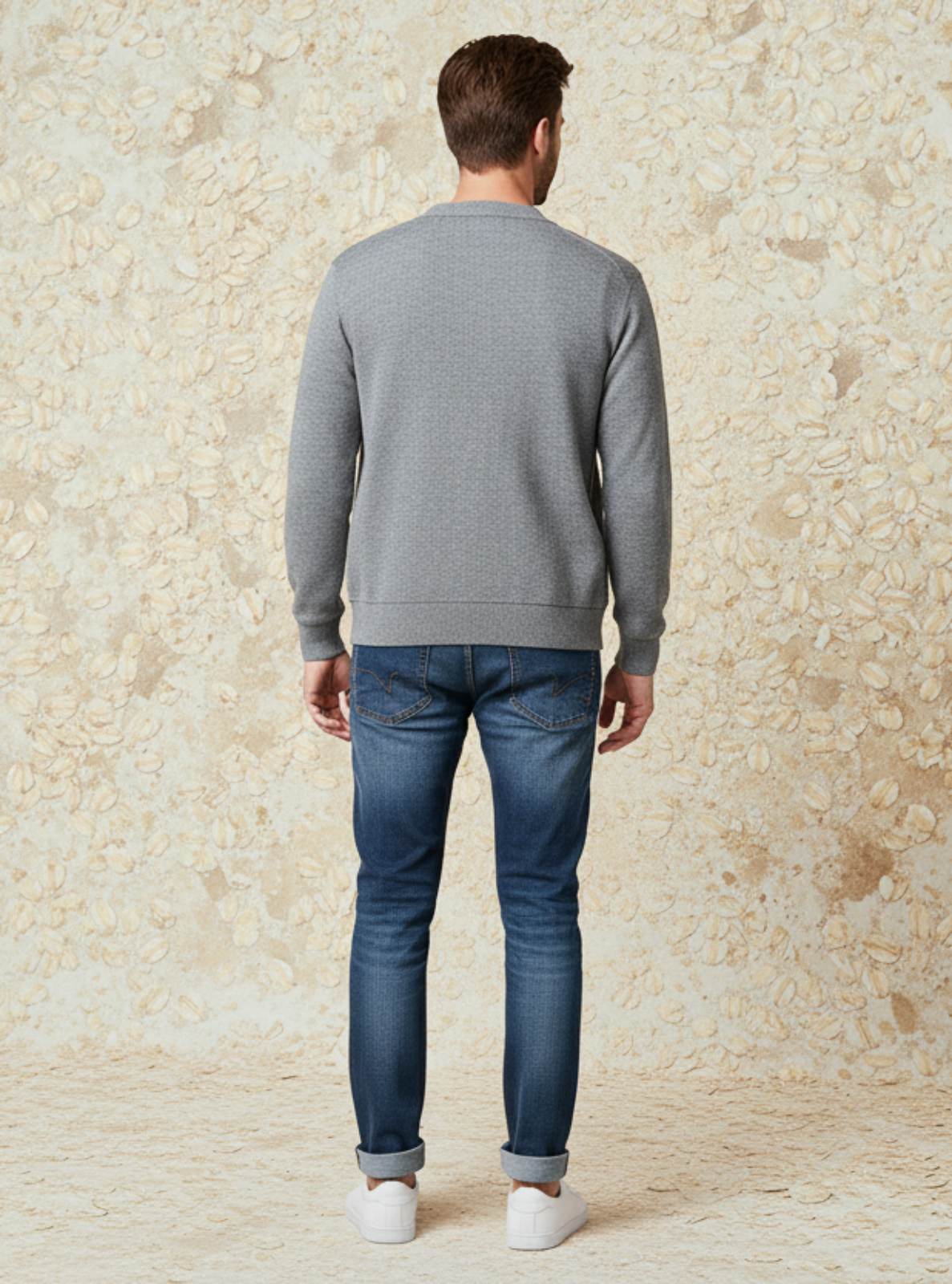 Man wearing a gray sweater and blue jeans standing against a textured beige wall.