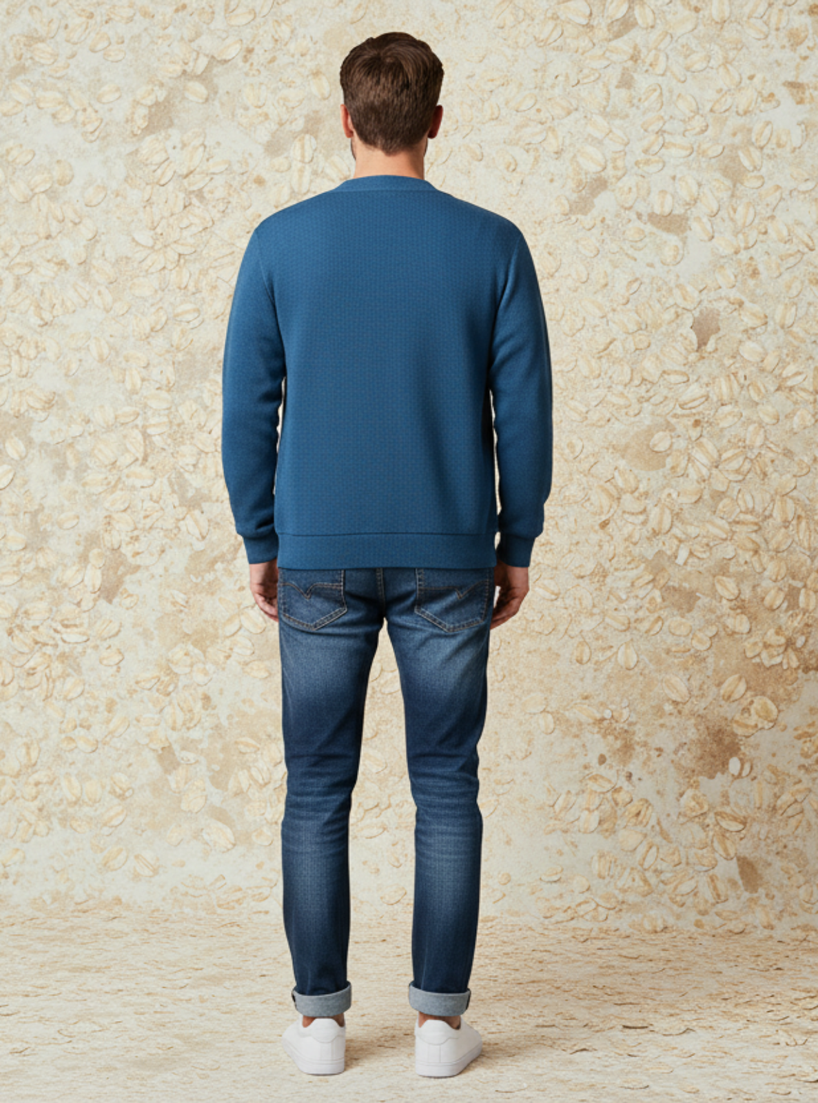 Man wearing a blue sweatshirt and jeans standing against a textured beige wall.