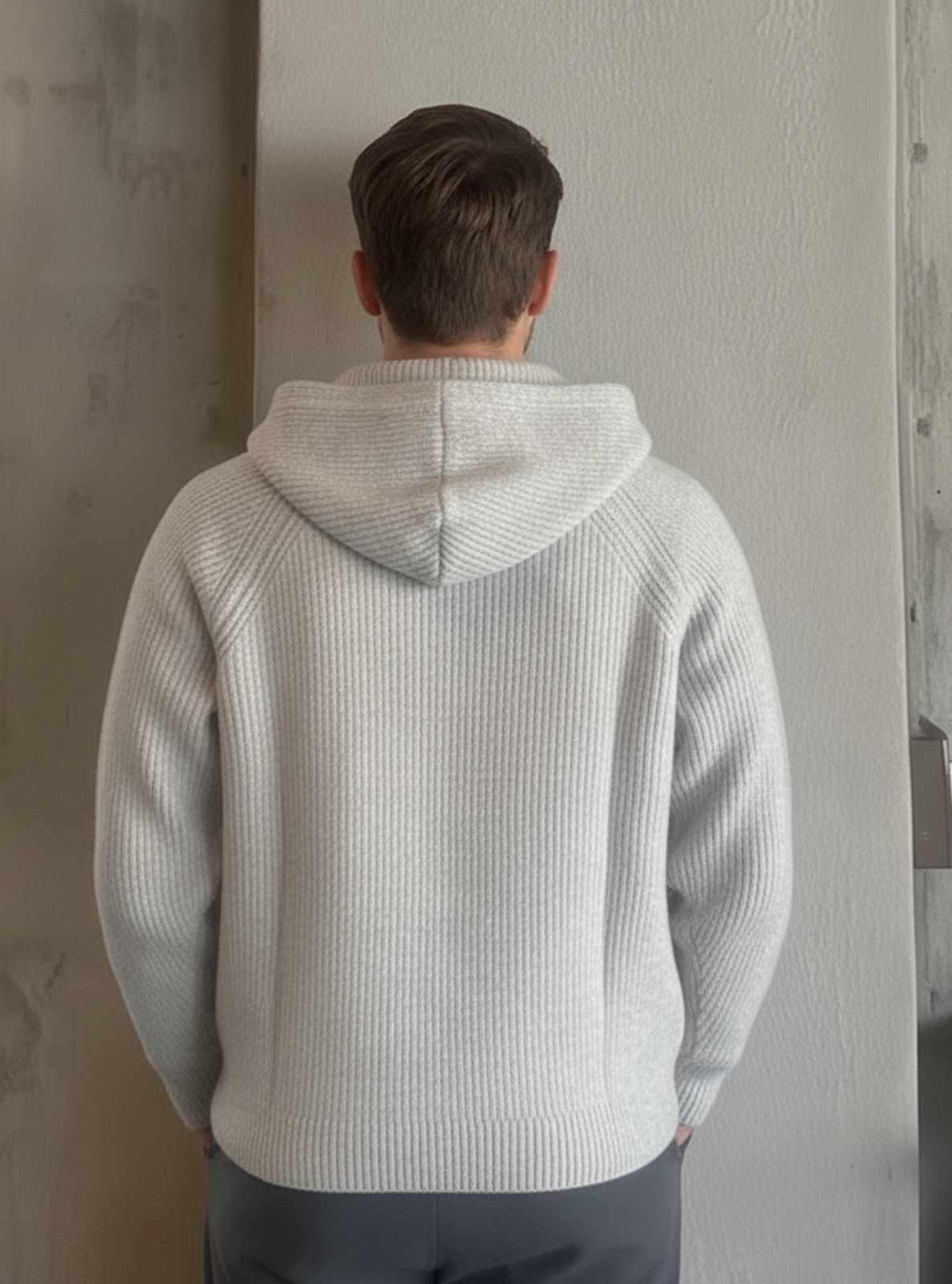 Person wearing a light gray hoodie standing against a wall indoors.