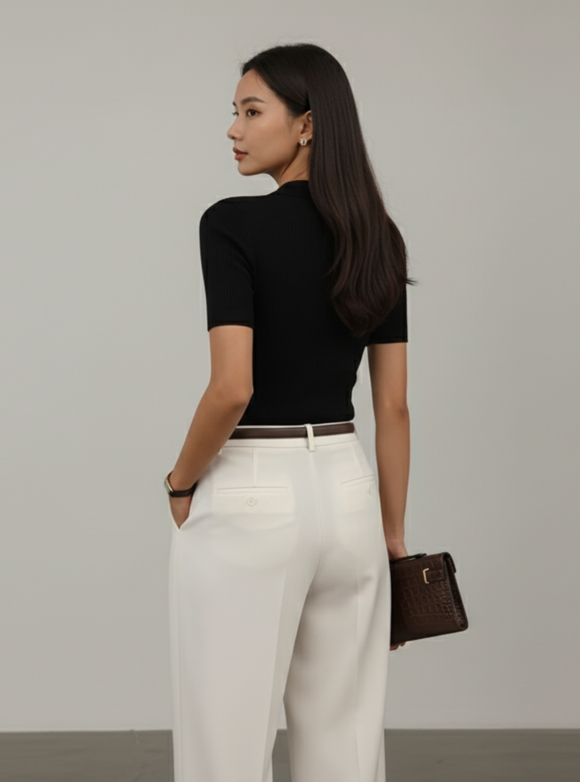 Woman wearing a black top and white pants with a brown handbag on a plain background