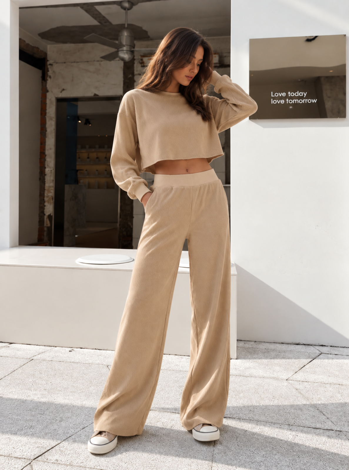 A woman modeling a beige two-piece lounge set consisting of a cropped long-sleeve top and matching wide-leg pants, standing on a stone patio in front of a white building.