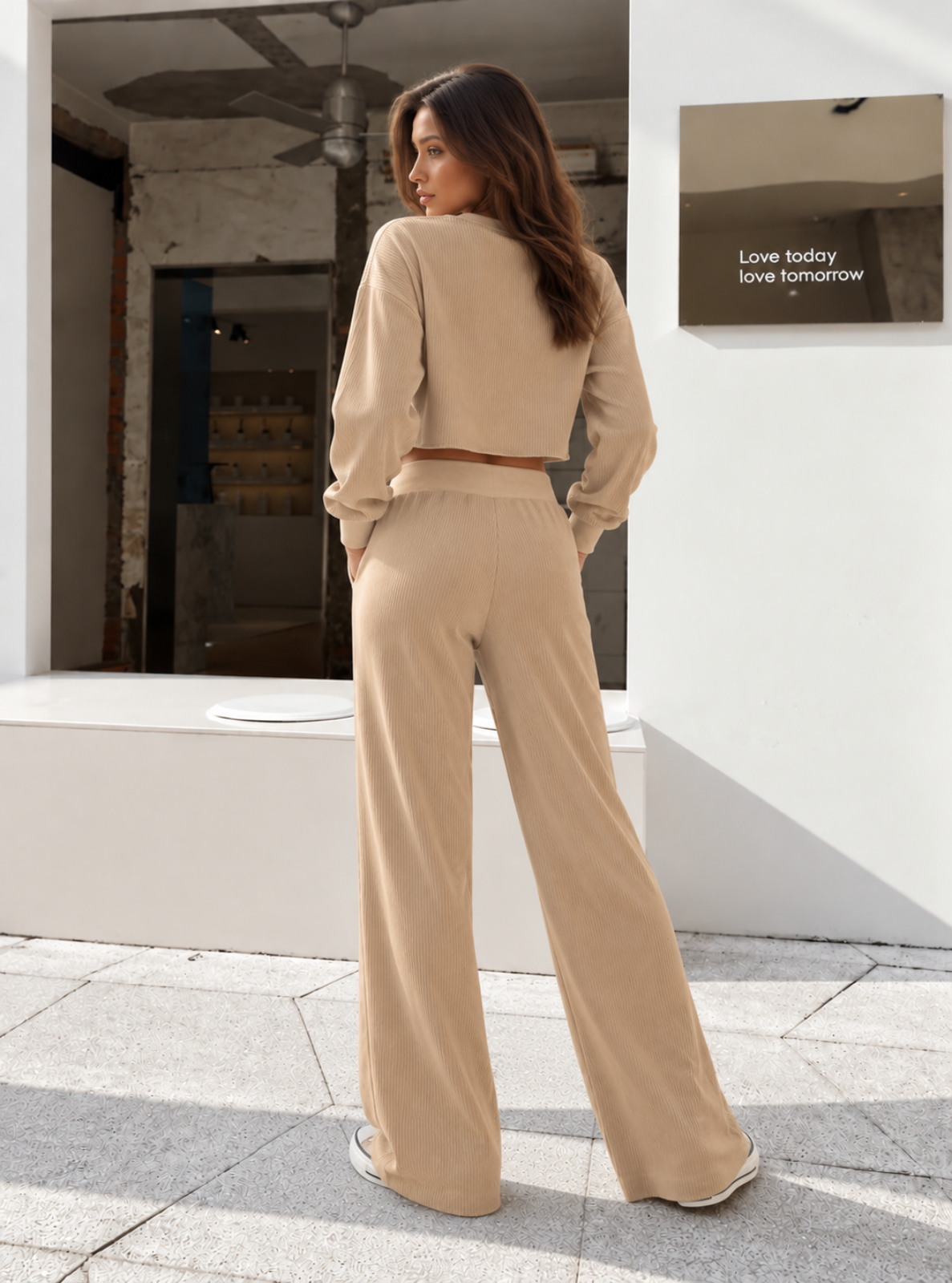 A woman wearing a beige two-piece lounge set consisting of a cropped long-sleeve top and matching wide-leg pants, standing outdoors in front of a white wall.
