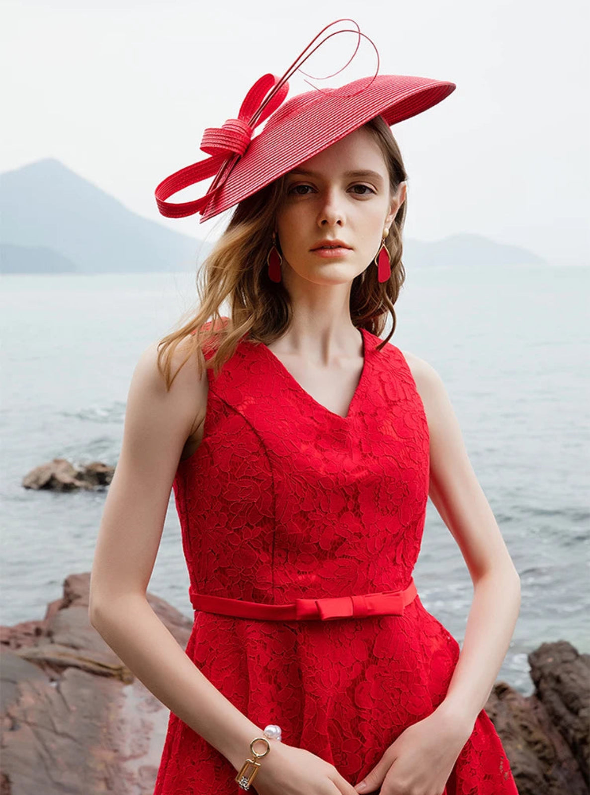 Woman in a red dress and hat standing by a body of water with mountains in the background