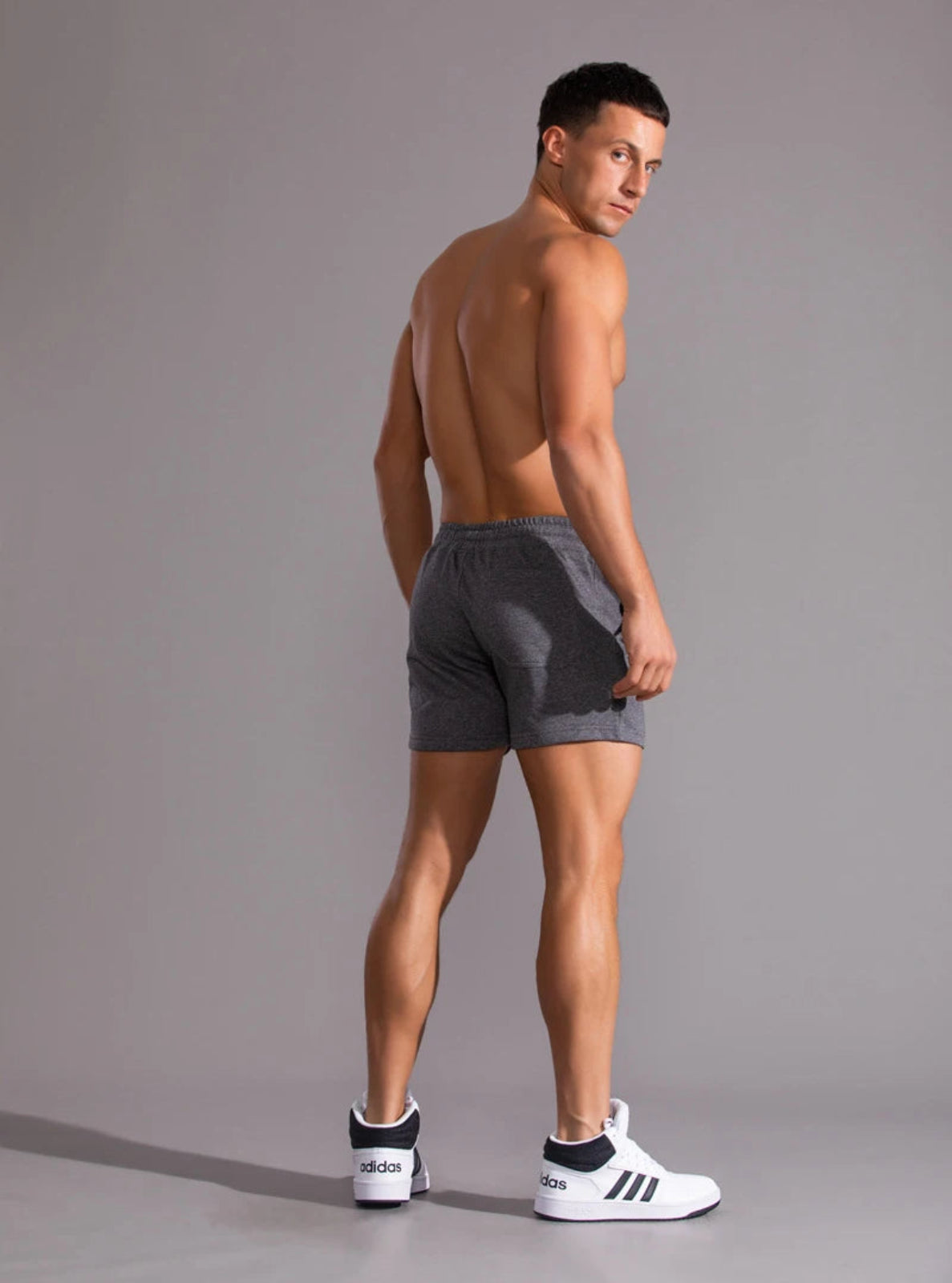 Man wearing gray athletic shorts and white sneakers with black stripes on a plain background