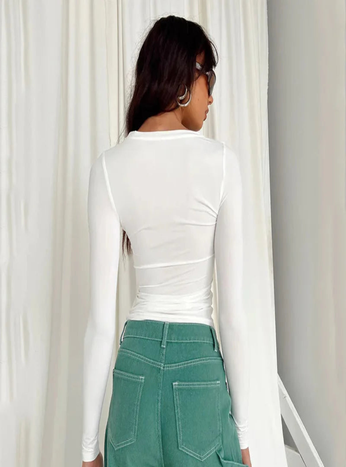 Person wearing a white long-sleeve top and green pants standing in front of white curtains.