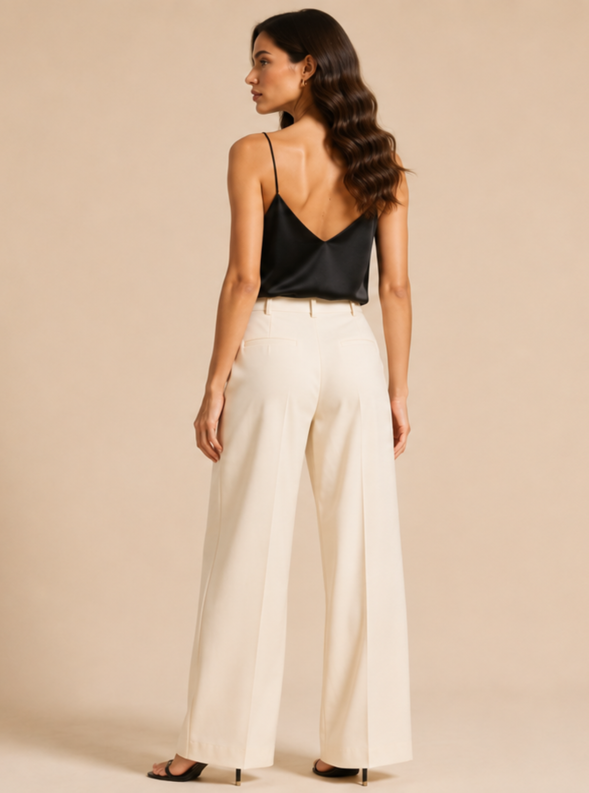 A woman is shown from behind wearing a black spaghetti-strap camisole top and high-waisted, wide-leg cream trousers, standing against a solid beige studio background.