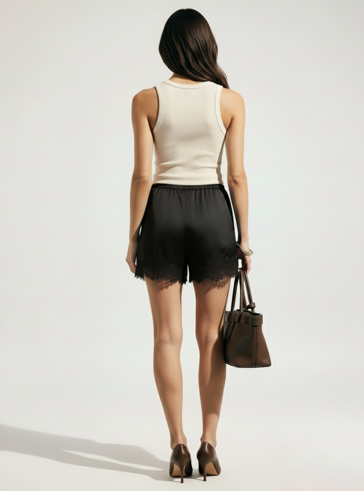 Woman wearing a beige sleeveless top and black shorts holding a brown handbag on a white background