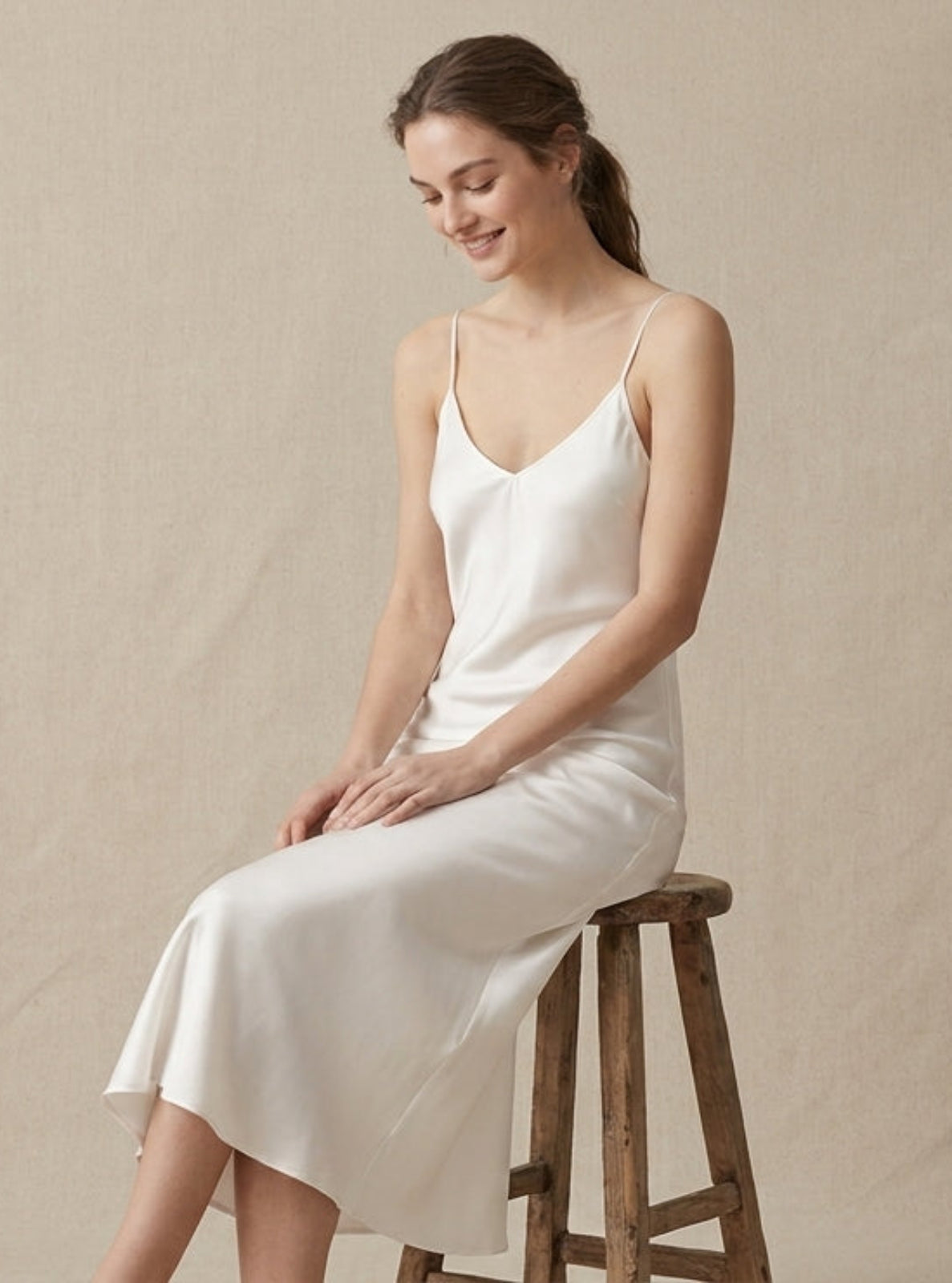 A woman wearing a white satin slip dress sits on a rustic wooden stool against a neutral beige textured backdrop.