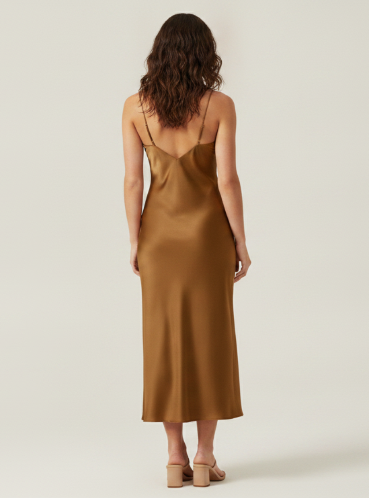 A woman viewed from behind wearing a satin slip dress in a warm camel brown color, standing against a plain light beige studio background.