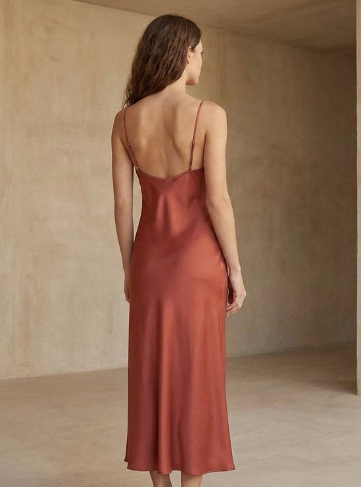 A woman viewed from behind wearing a rust-colored satin slip dress with thin spaghetti straps and a low open back, set against a warm, textured beige plaster wall and floor.