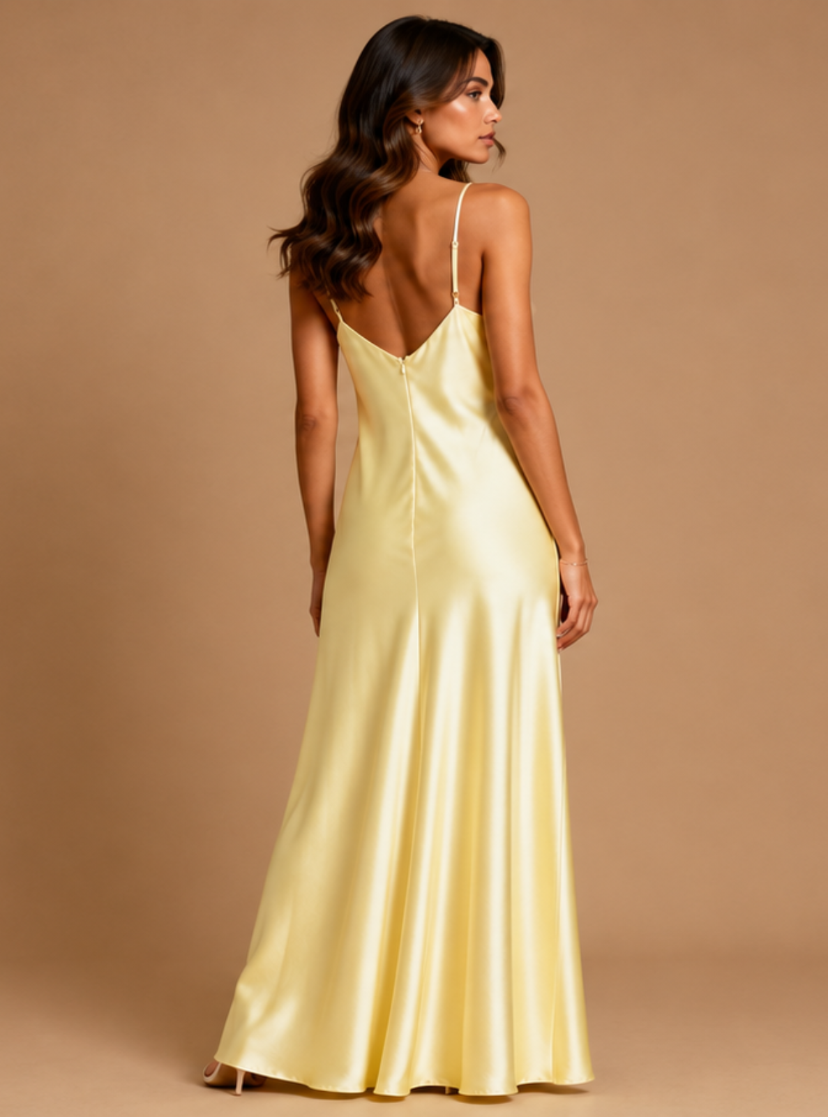 A woman modeling a floor-length satin slip dress from the back, shown against a solid warm tan studio background.