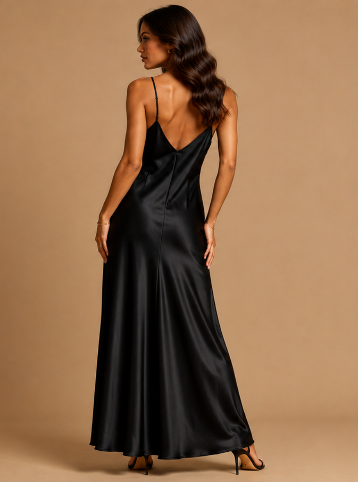 A woman modeling a black satin maxi dress from the back, featuring thin spaghetti straps, a deep V-back, and a flowing floor-length silhouette, set against a solid warm beige studio background.