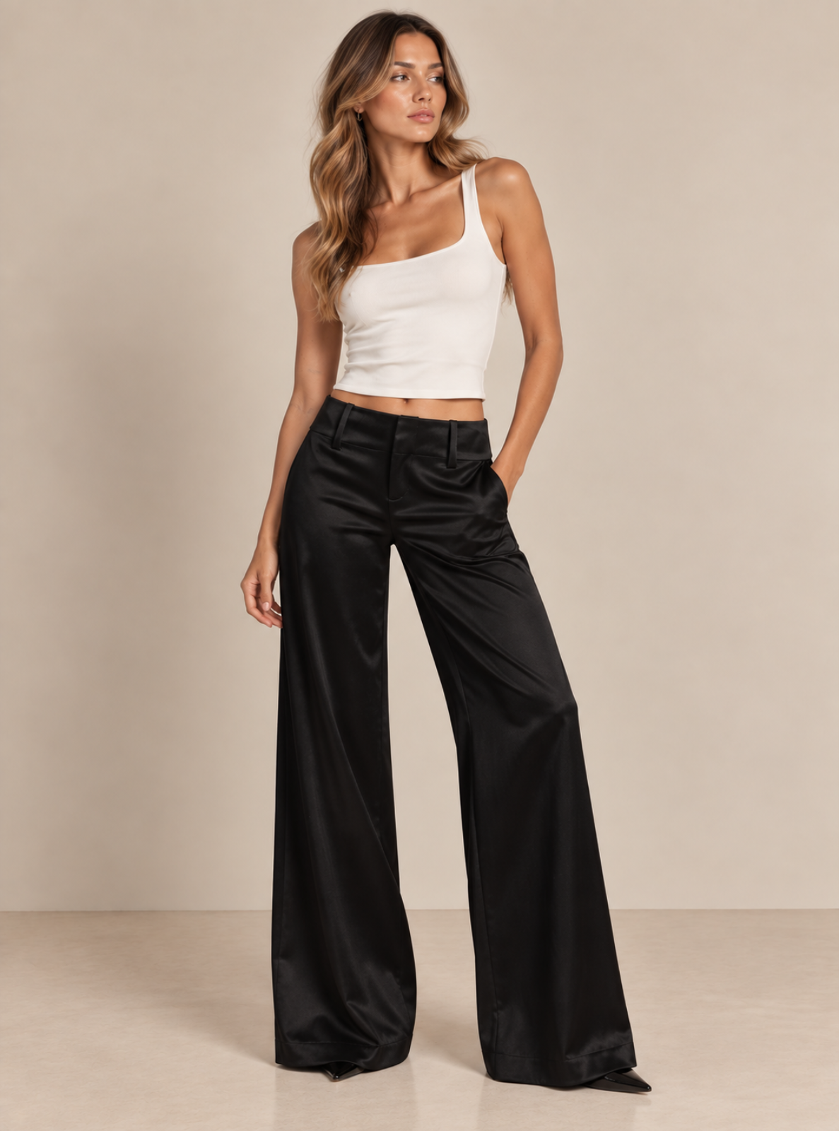 A woman modeling black wide-leg satin pants and a white one-shoulder crop top against a plain beige studio backdrop.