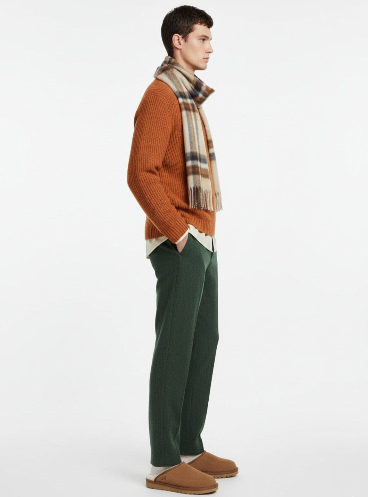 Man wearing an orange sweater, plaid scarf, green pants, and brown shoes on a white background