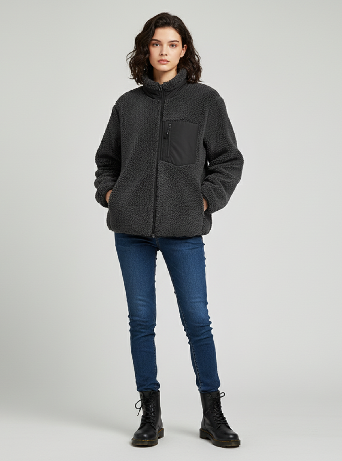 Person wearing a dark gray fleece jacket, blue jeans, and black boots on a light gray background