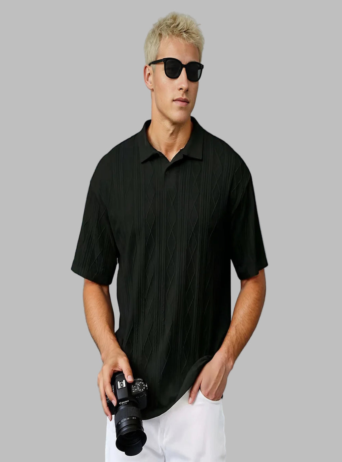 Man wearing a black textured polo shirt and sunglasses, holding a camera against a light blue wall.