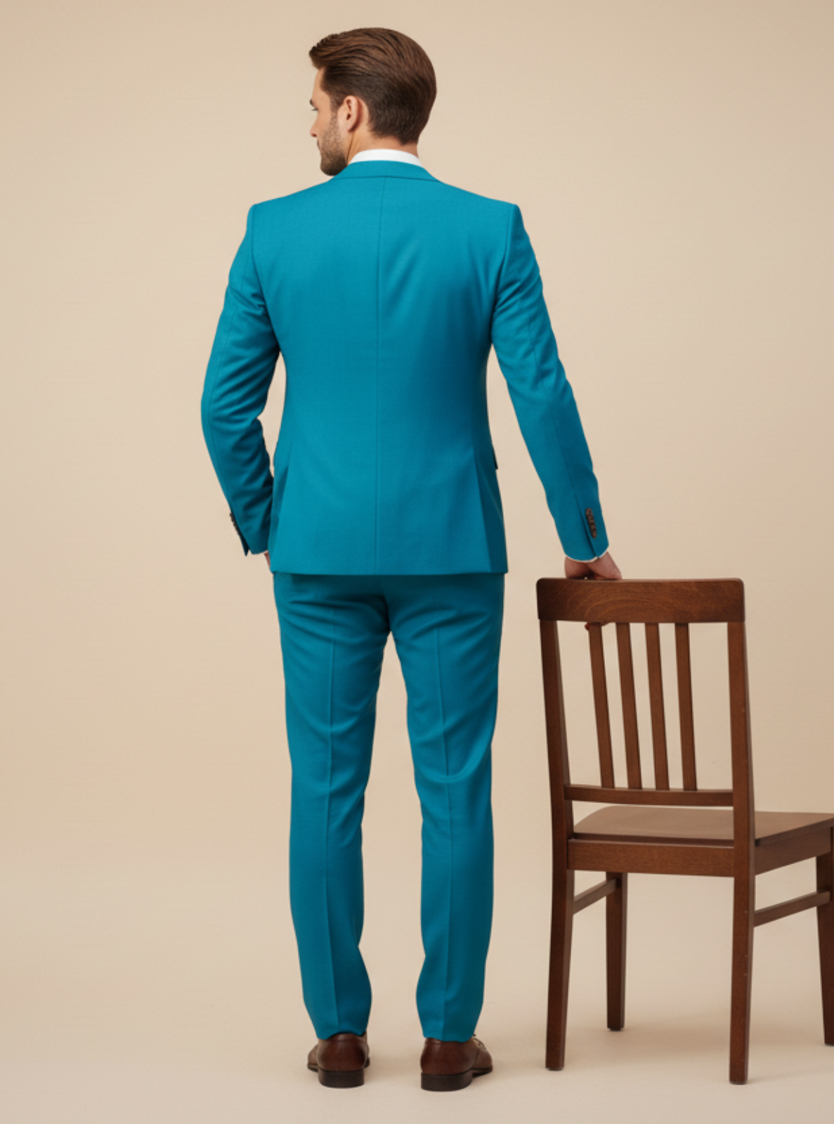 Man in a teal suit standing next to a wooden chair on a beige background