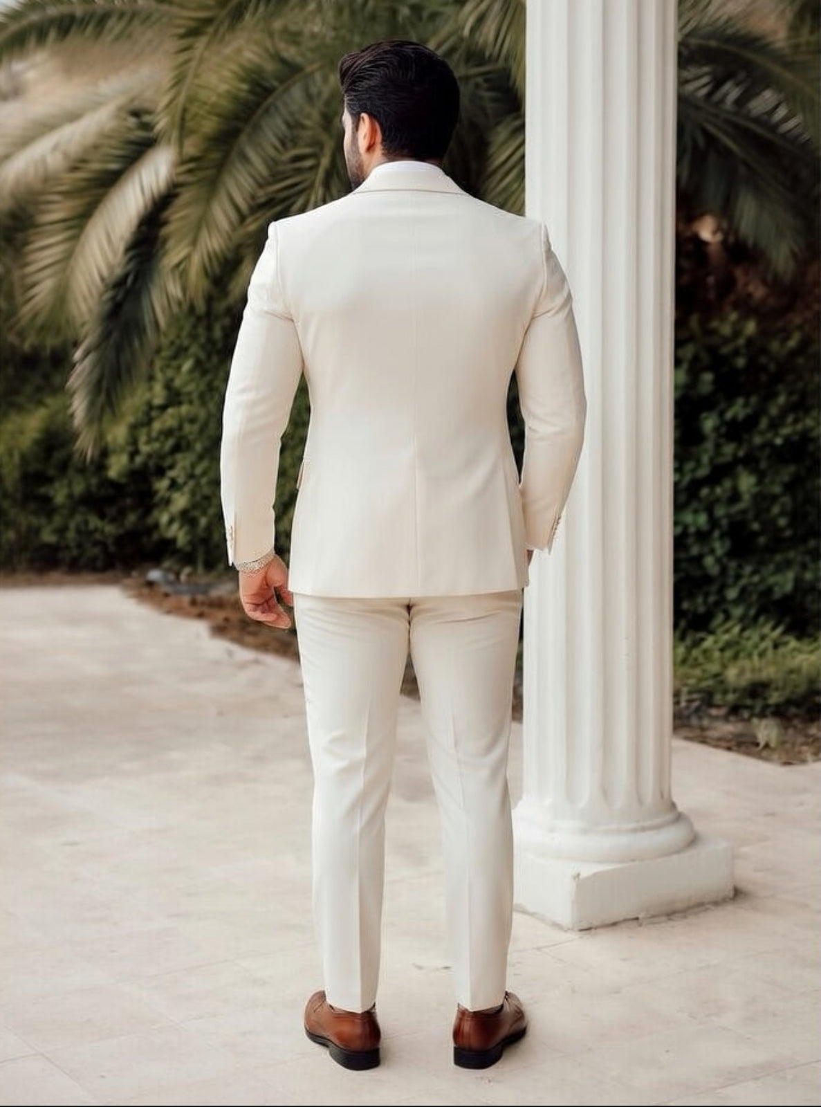 Man in a white suit standing in front of a classical column with palm trees in the background