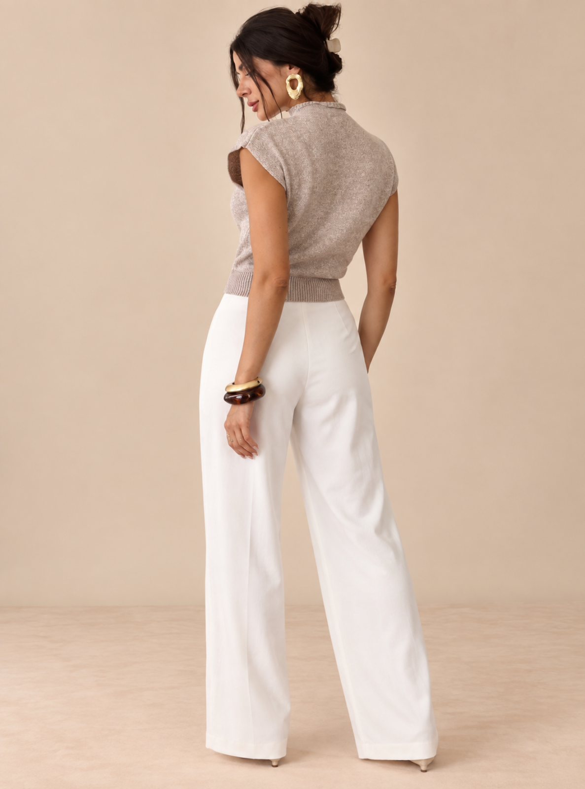 A woman shown from the back wearing a sleeveless taupe knit mock-neck top and white wide-leg pants, standing against a solid beige studio background.