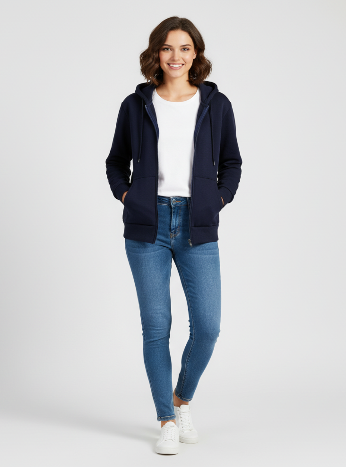 Woman wearing a navy hoodie, white t-shirt, blue jeans, and white sneakers on a light gray background
