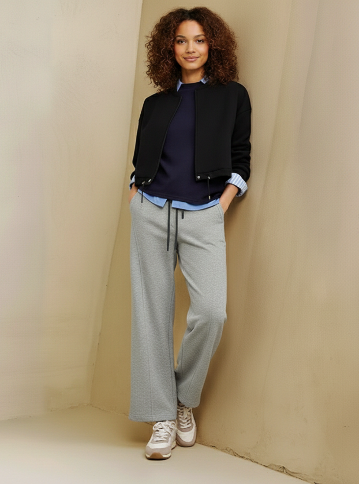 Woman wearing a black jacket, navy shirt, and gray pants against a beige wall.