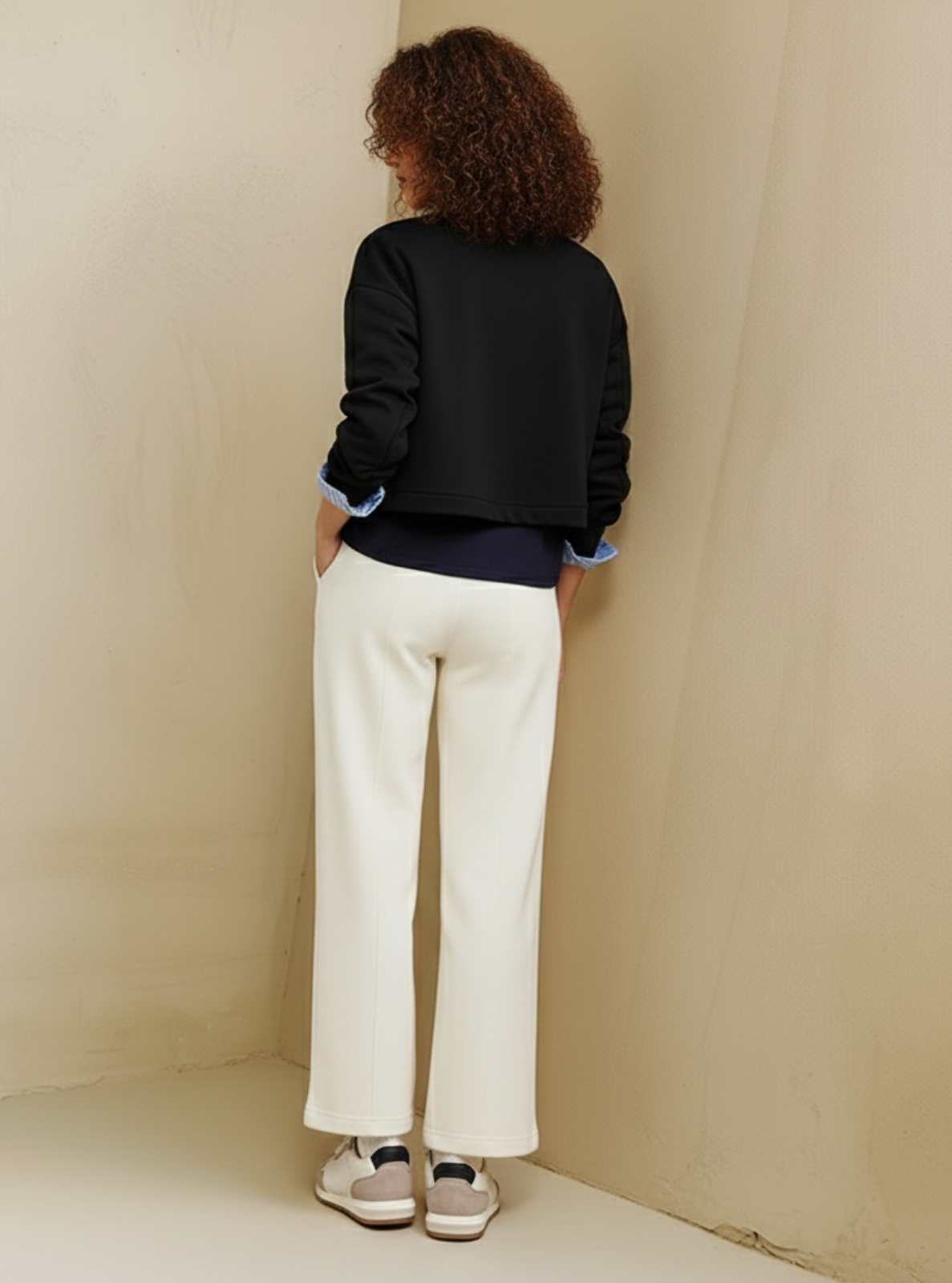 Person wearing a black jacket and white pants standing in a corner of a room with beige walls.
